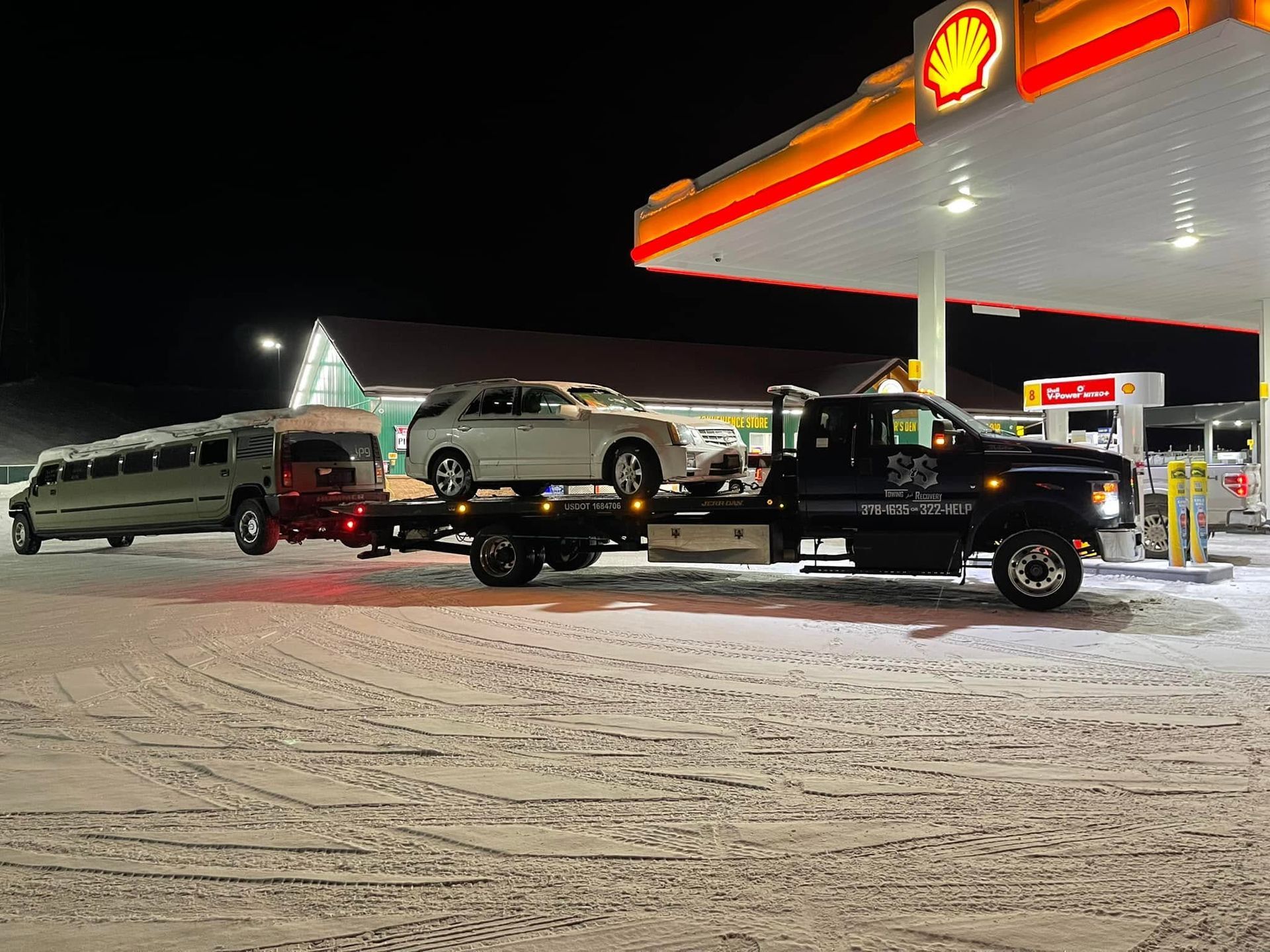 Cars on a tow truck outside a Shell gas station at night with snow on the ground