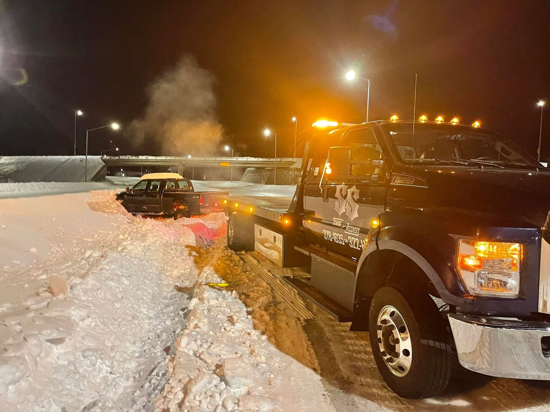 Tow truck pulling a car from deep snow at night in a snowy parking lot