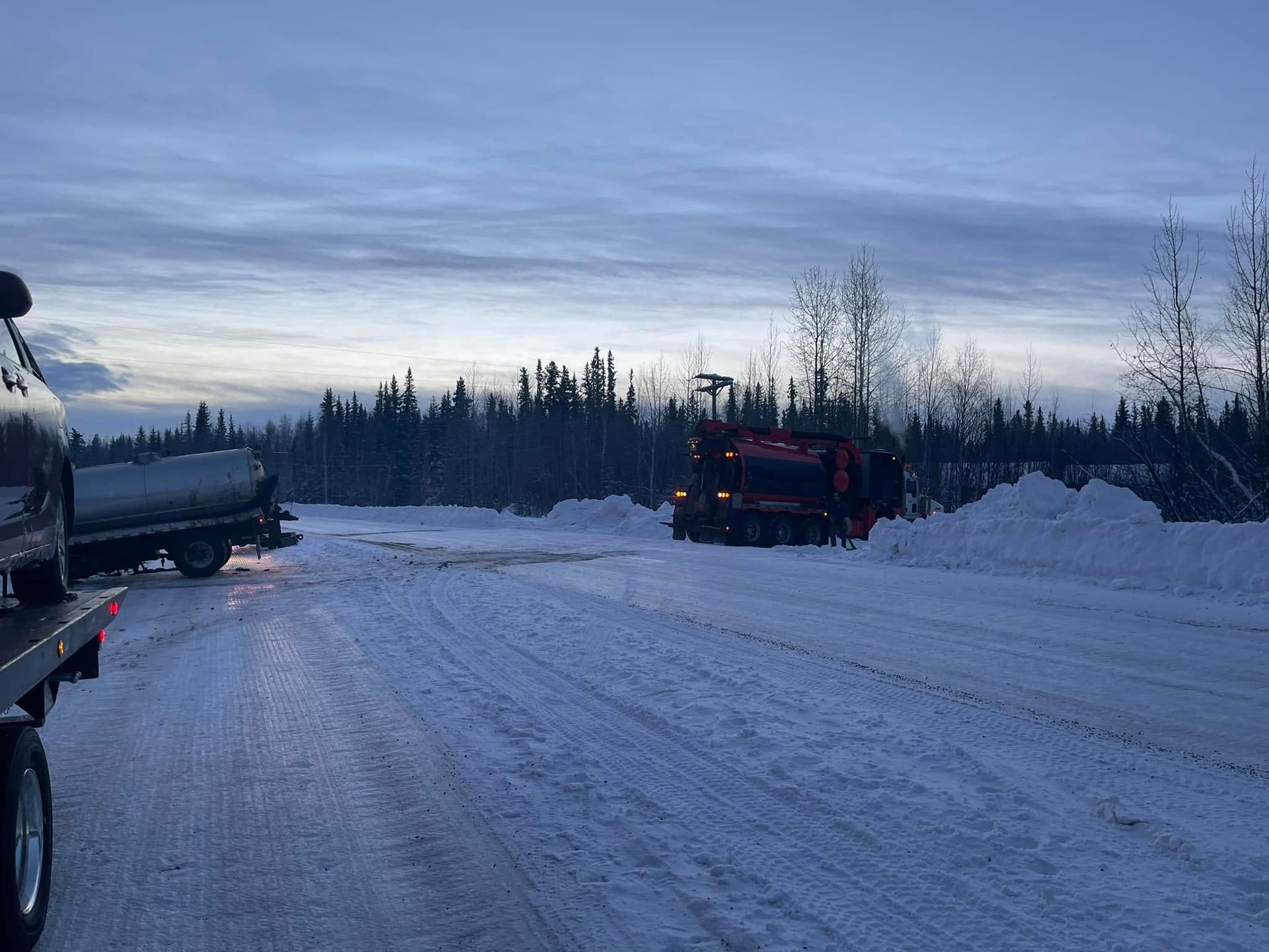 Snow-covered roadside at dusk with a truck parked on the left and a red vehicle ahead near a forest.