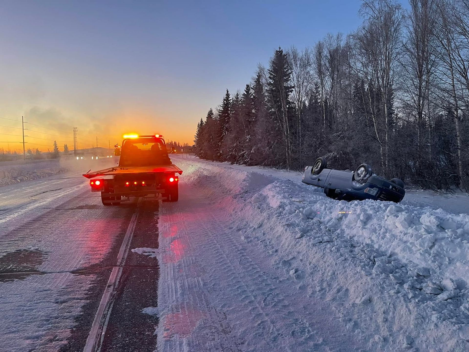Tow truck towing a car on a snowy road at sunset, with snowbanks and trees along the roadside