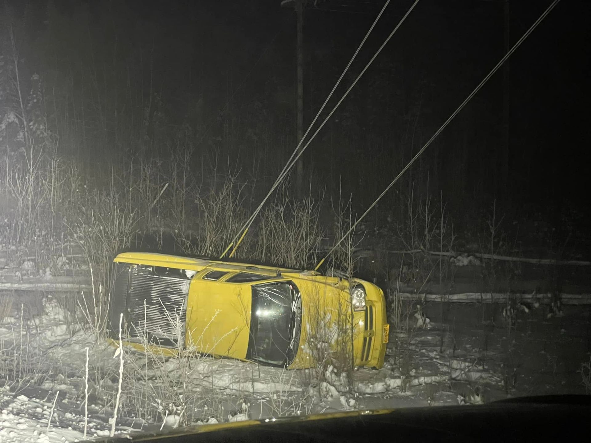Yellow car overturned in snow at night, with power lines overhead.