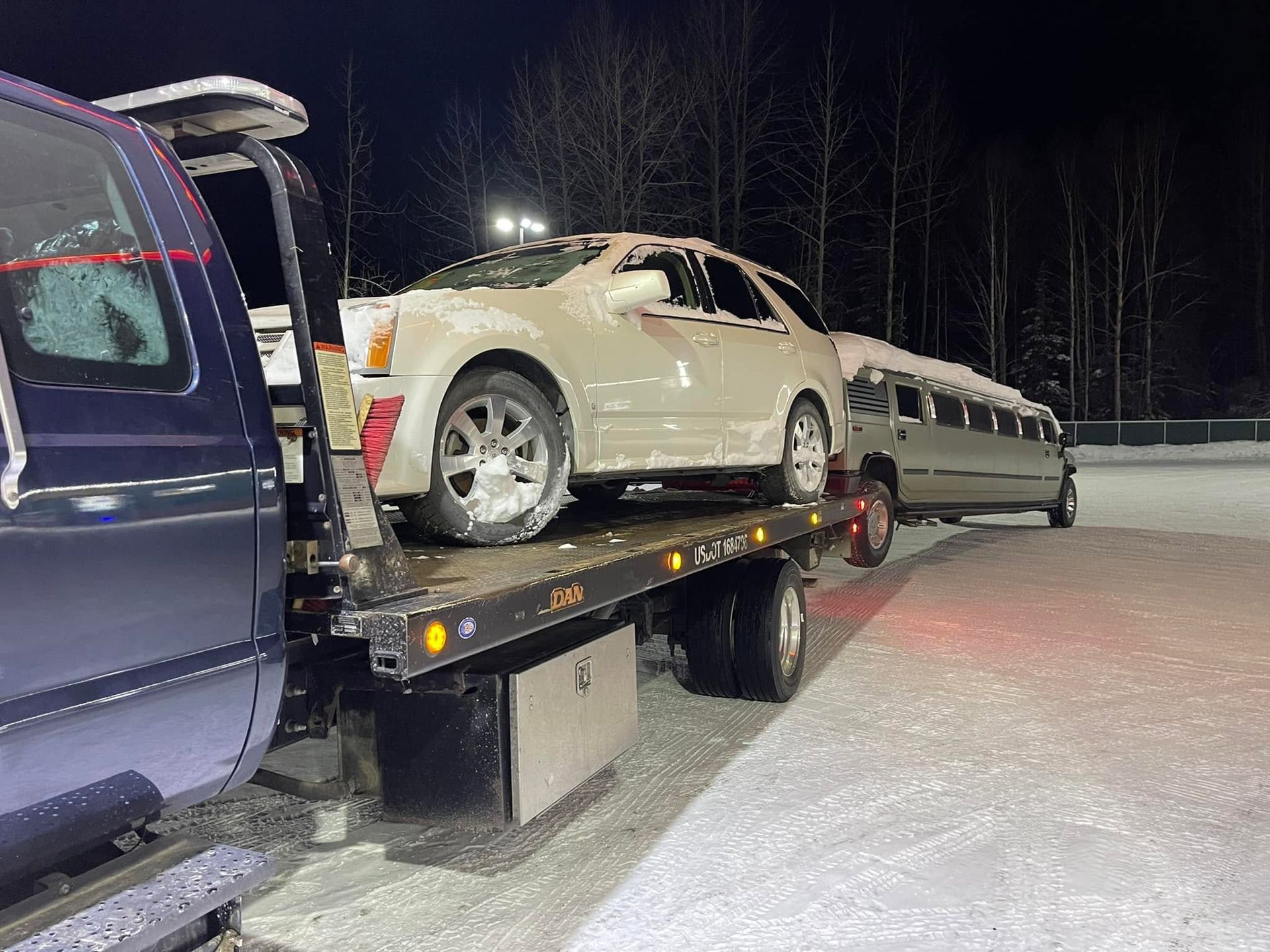 Car on a tow truck at night in a snowy lot, with a van beside it.