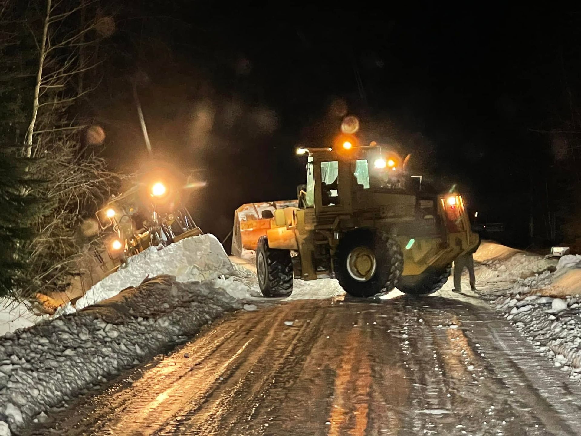 Snowplow clearing a snowy road at night, with bright lights and piled snowbanks on both sides