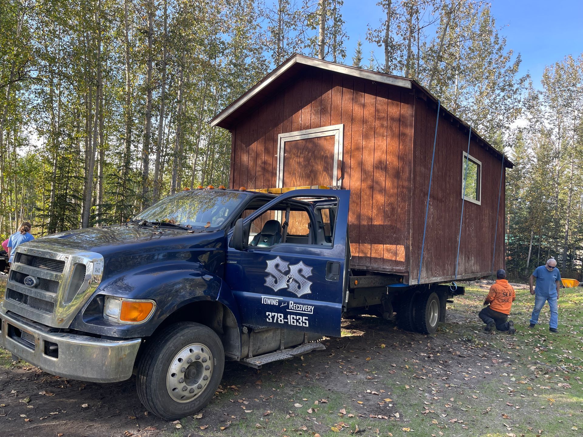 Blue truck hauling a small brown cabin in a wooded clearing, with two people nearby
