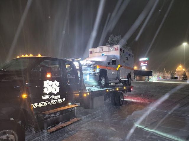 Tow truck carrying a white pickup on a snowy night road with bright lights and blowing snow