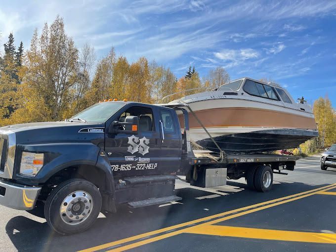 Tow truck hauling a white and black boat on a trailer along a road with autumn trees.