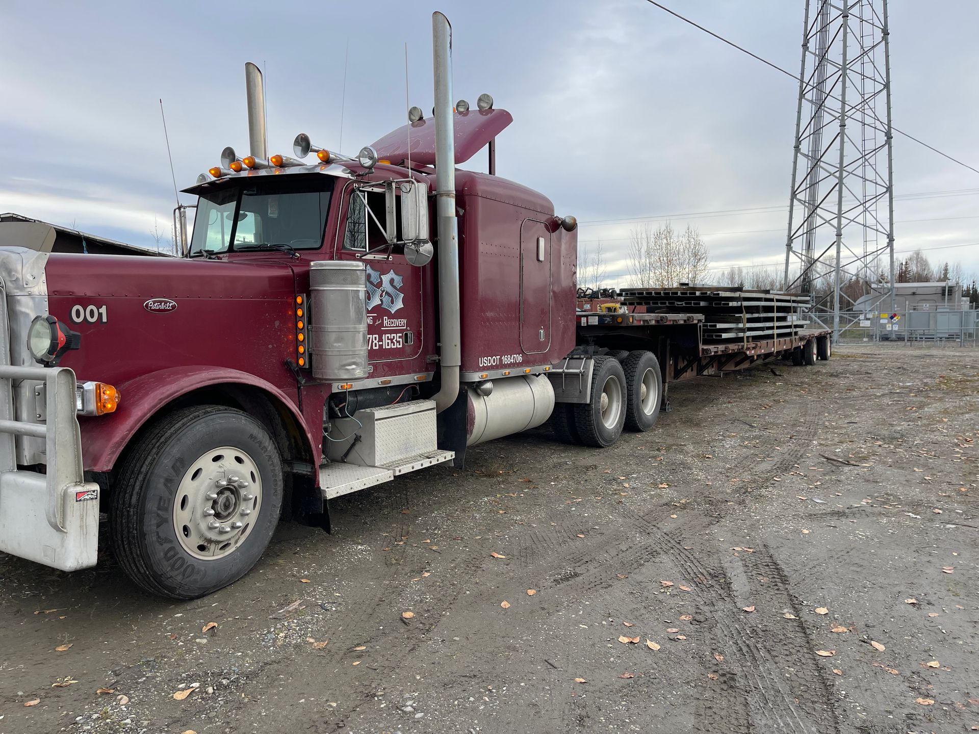 Maroon semi-truck hauling stacked metal equipment on a gravel lot under cloudy skies