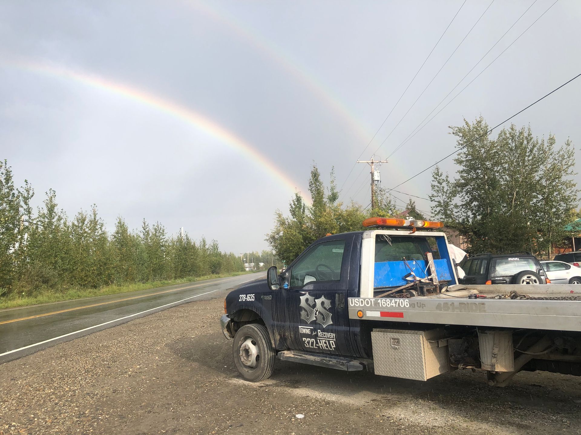 Tow truck on a wet roadside under a double rainbow, with trees and parked cars nearby