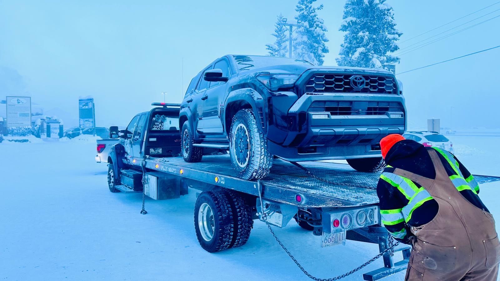 Tow truck hauling two pickup trucks on a snowy road, with a worker securing the load
