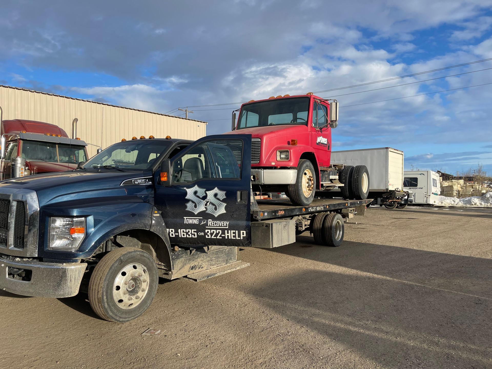 Black flatbed tow truck carrying a red truck in an industrial lot under a cloudy sky