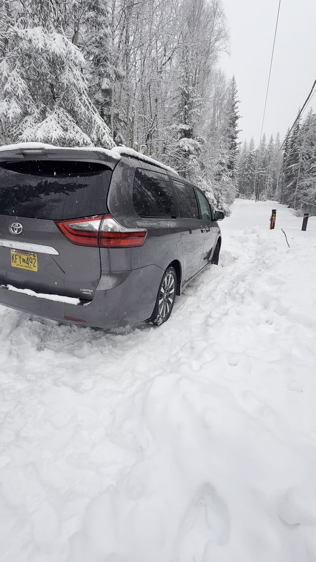 Gray minivan stuck in deep snow on a forest road, with snow-covered trees all around.
