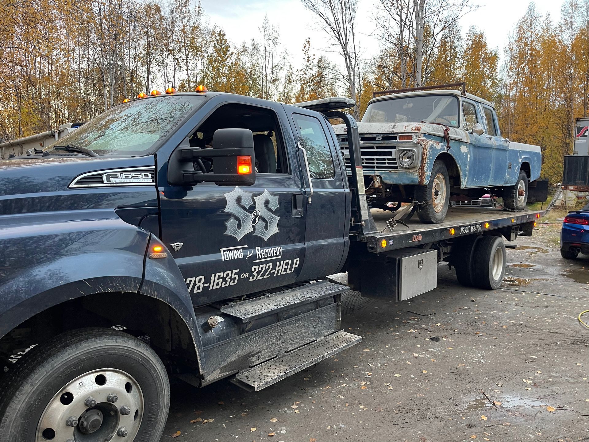 Blue tow truck hauling a rusted blue pickup on a flatbed in a gravel lot with autumn trees.