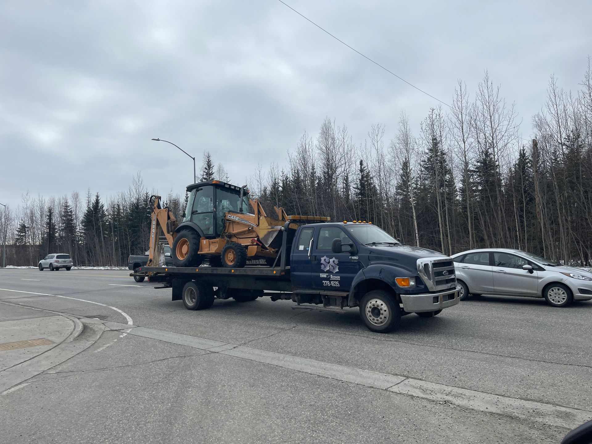 Tow truck carrying a backhoe on a road beside a silver car, under a cloudy sky