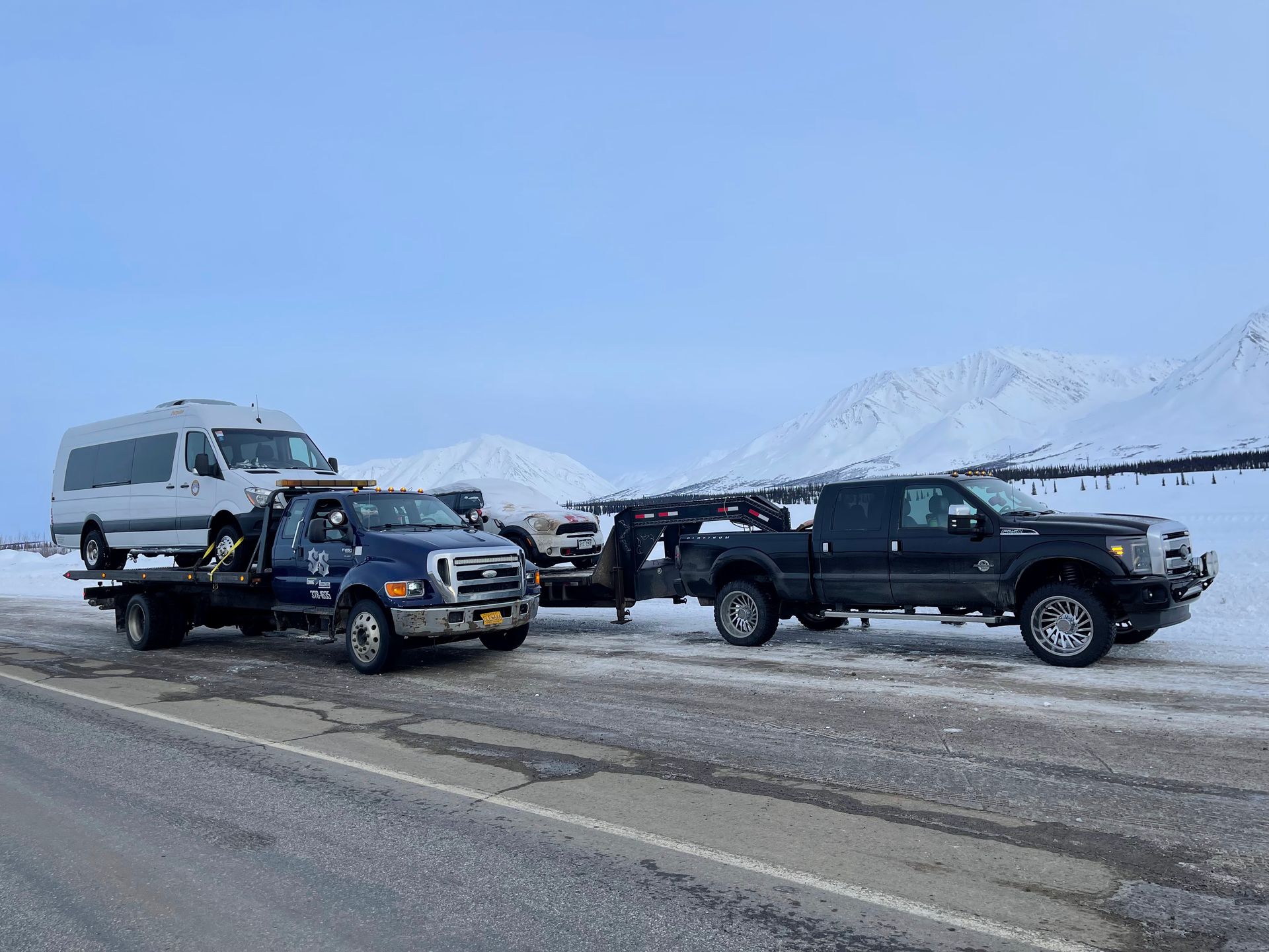 Two pickup trucks towing a van on a snowy roadside with mountains in the background