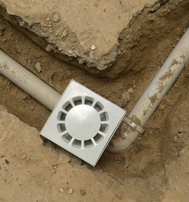 A square white drainage grate connected to two white pipes in a dirt trench.