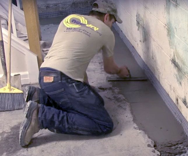 Worker kneeling to smooth wet gray concrete along a wall inside a room