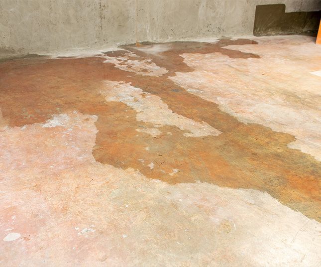 Stained concrete floor with large patches of peeling, discolored surface near a wall