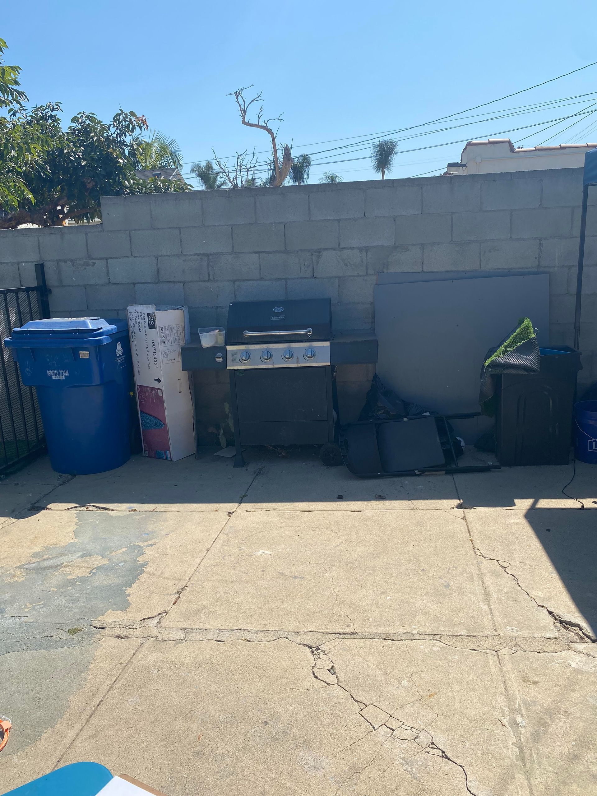 Backyard patio with a grill, trash bins, and a cinderblock wall under bright sunlight