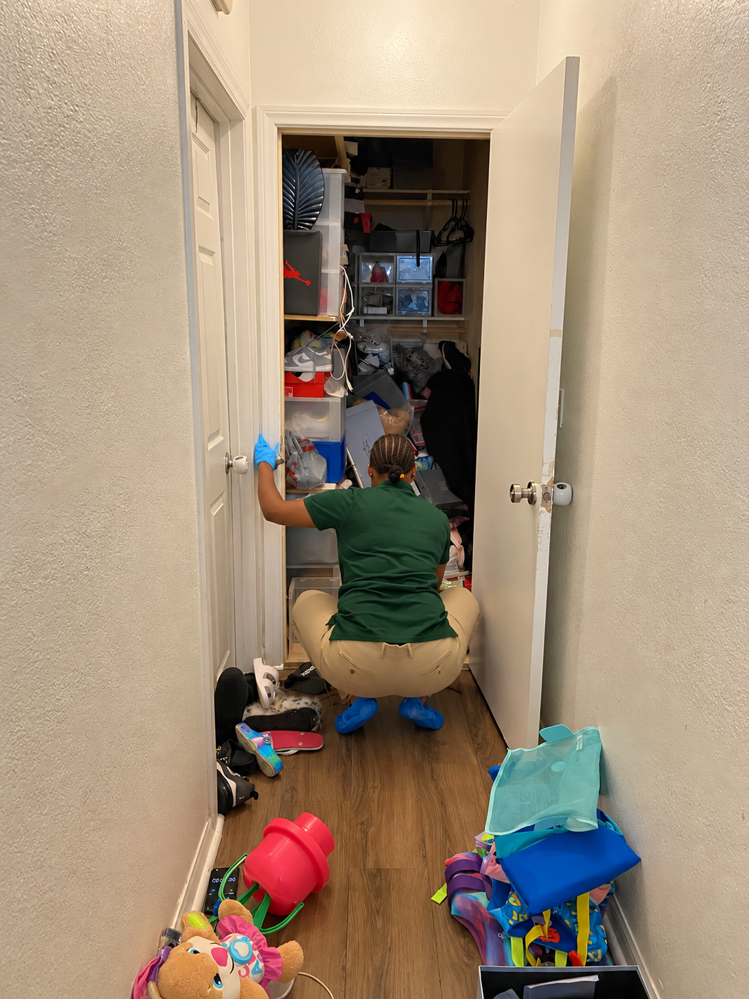 Person kneeling in a hallway, organizing a cluttered closet with bins and bags around them.