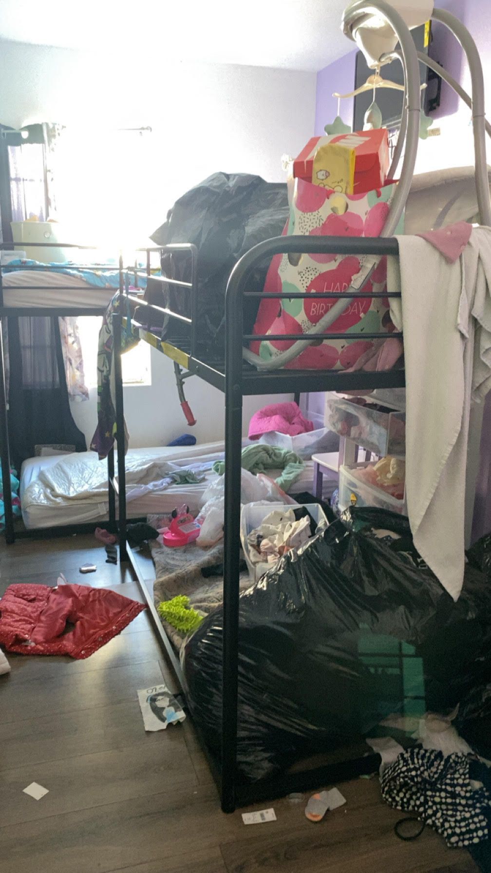 Cluttered bedroom with a metal clothing rack, piled bags, scattered clothes, and a bright window.
