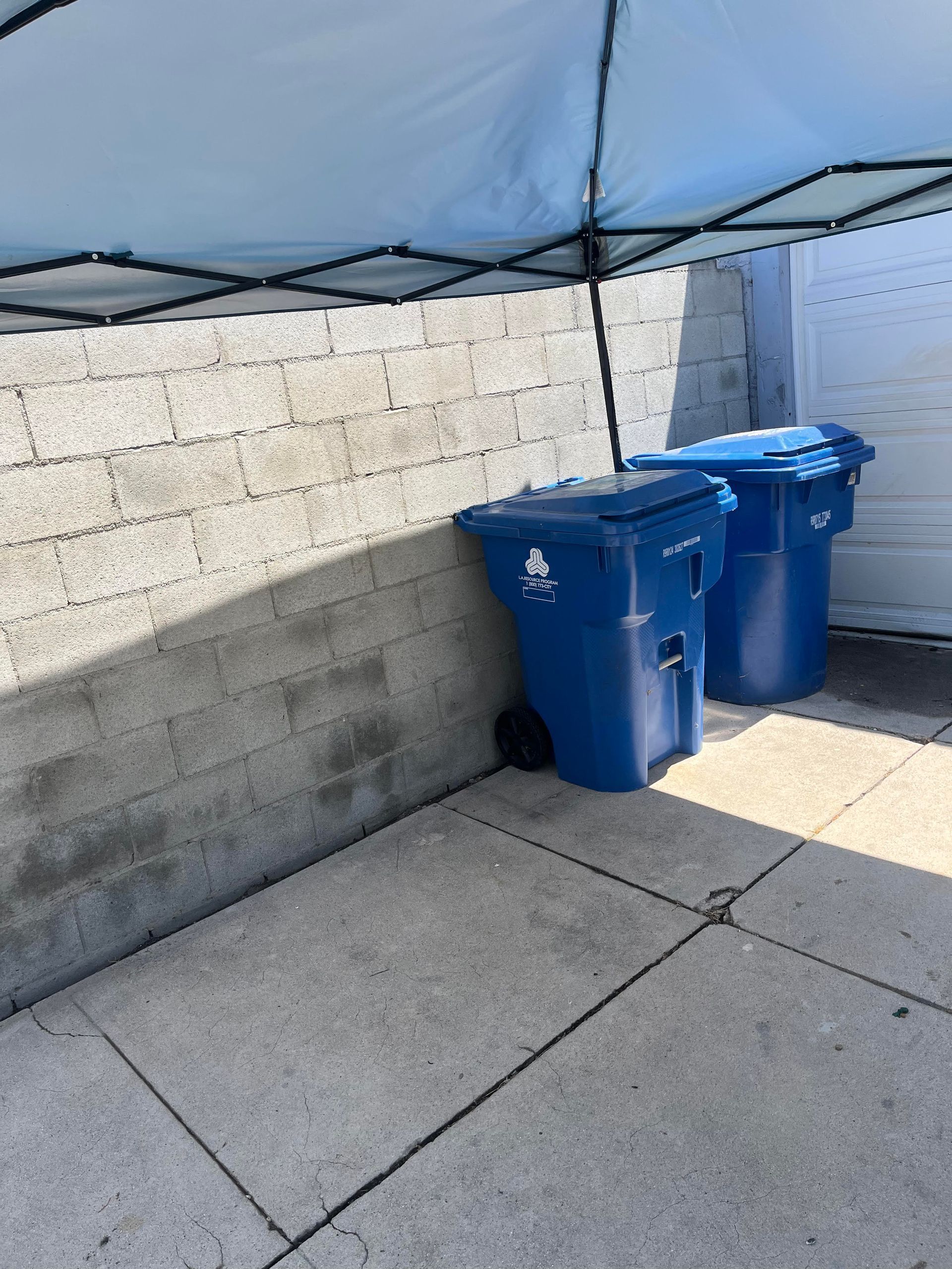 Two blue recycling bins beside a cinderblock wall under a blue canopy on a concrete patio