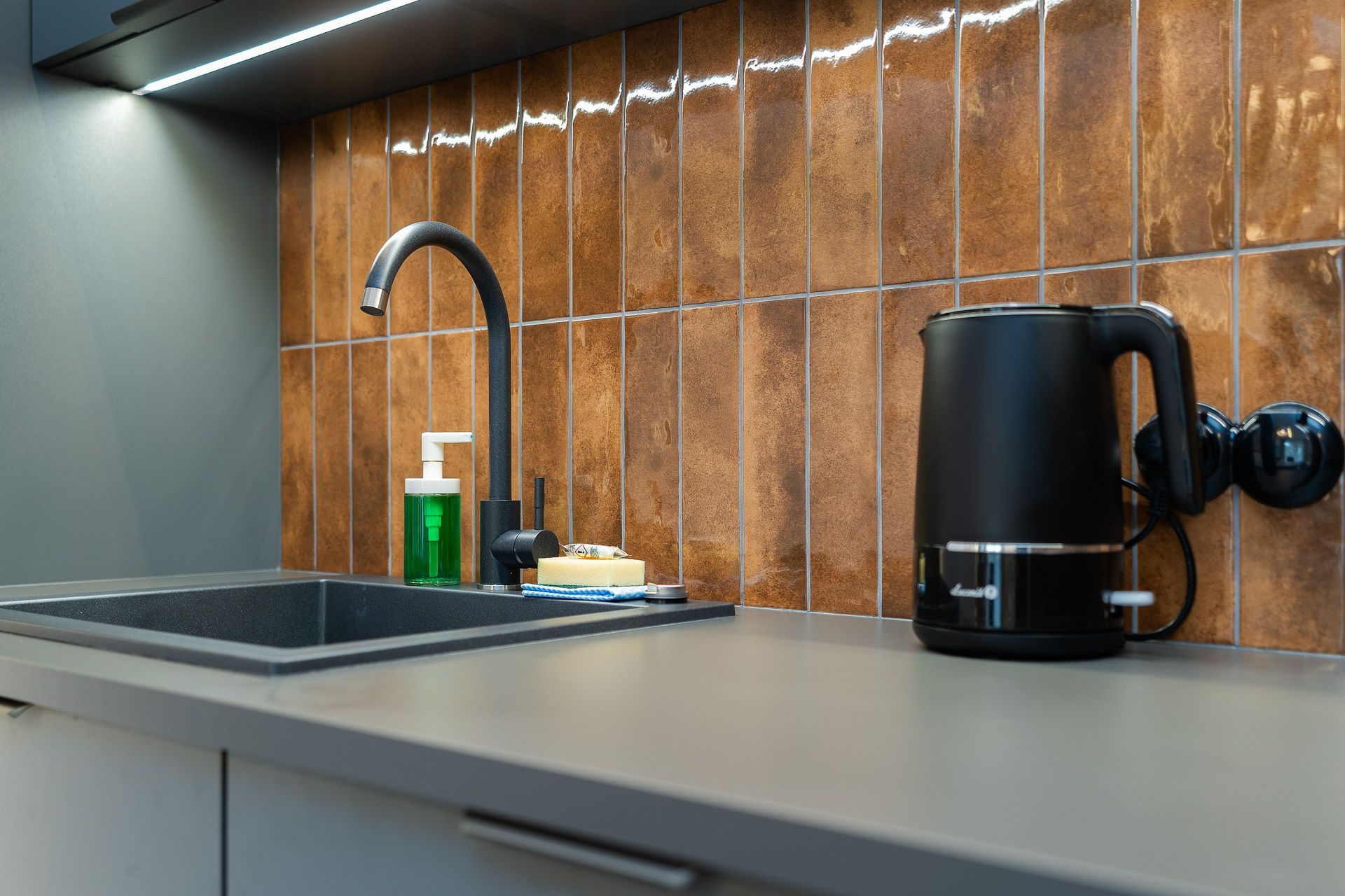 Modern kitchen counter with stainless sink, black faucet, soap dispenser, and black electric kettle under under-cabinet lighting