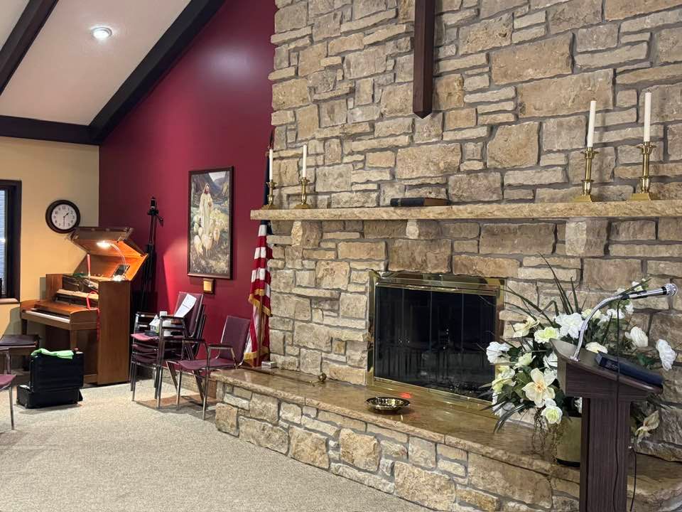 A church sanctuary featuring a stone fireplace, an upright piano, American flag, and floral arrangement on a pulpit.