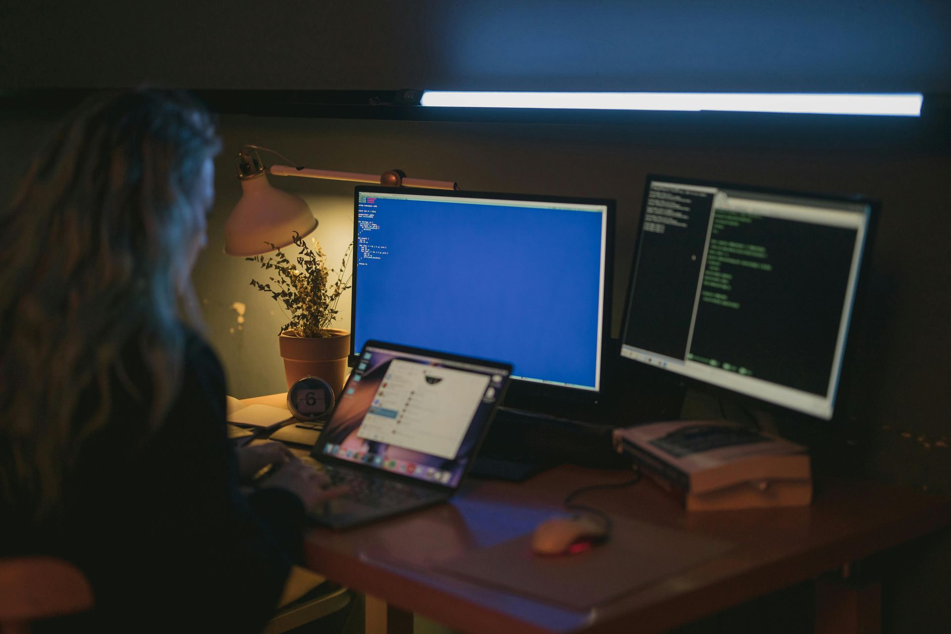 Woman at computer desk with multiple screens, coding in a dimly lit room.
