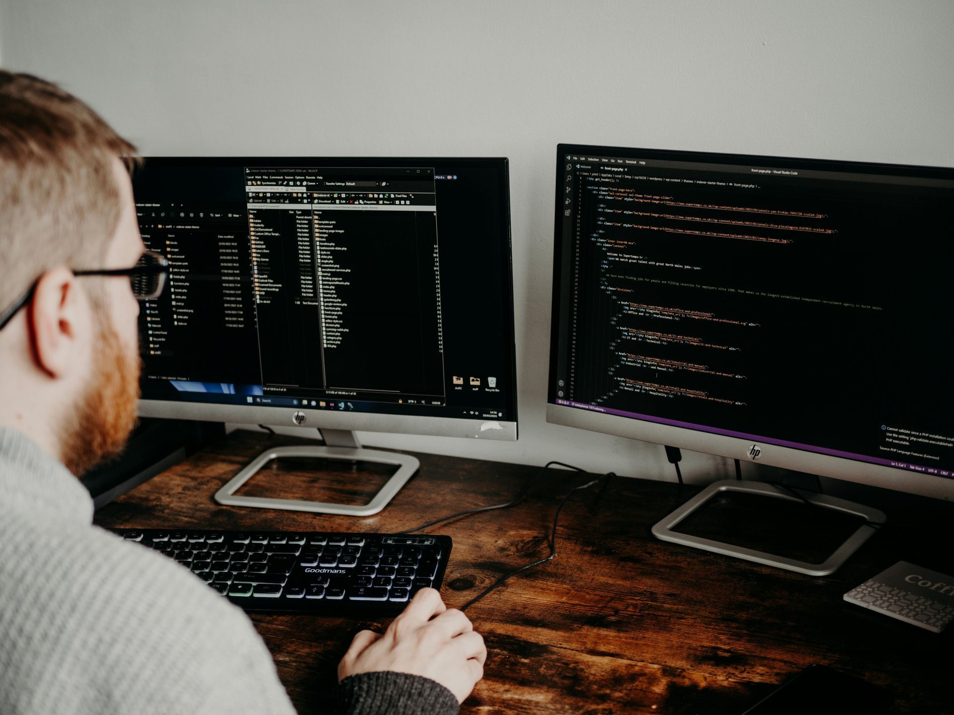 A person with glasses working on a computer with two monitors, displaying code, indoors.