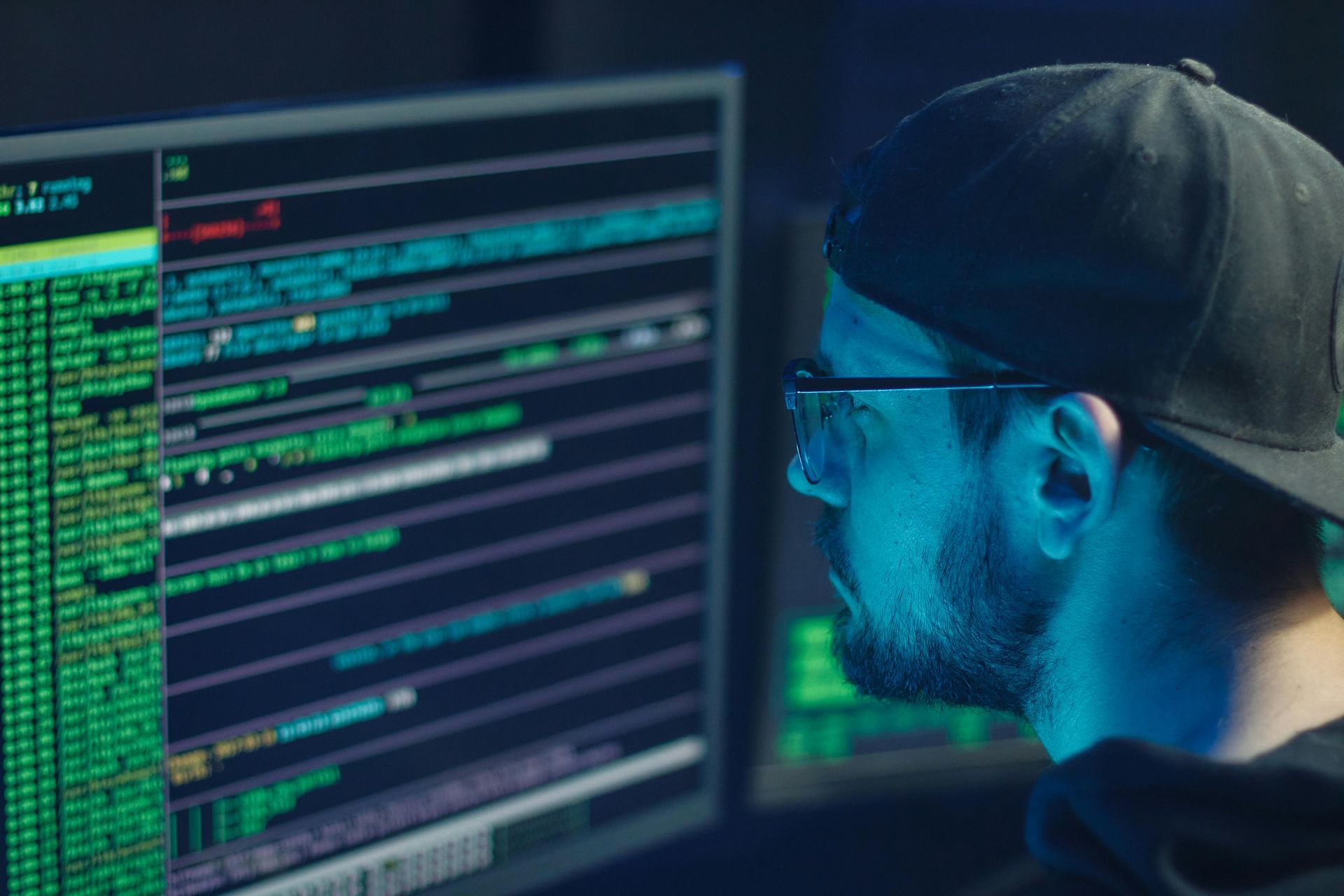 Man in cap and glasses, illuminated by blue light, analyzes code on computer screens.