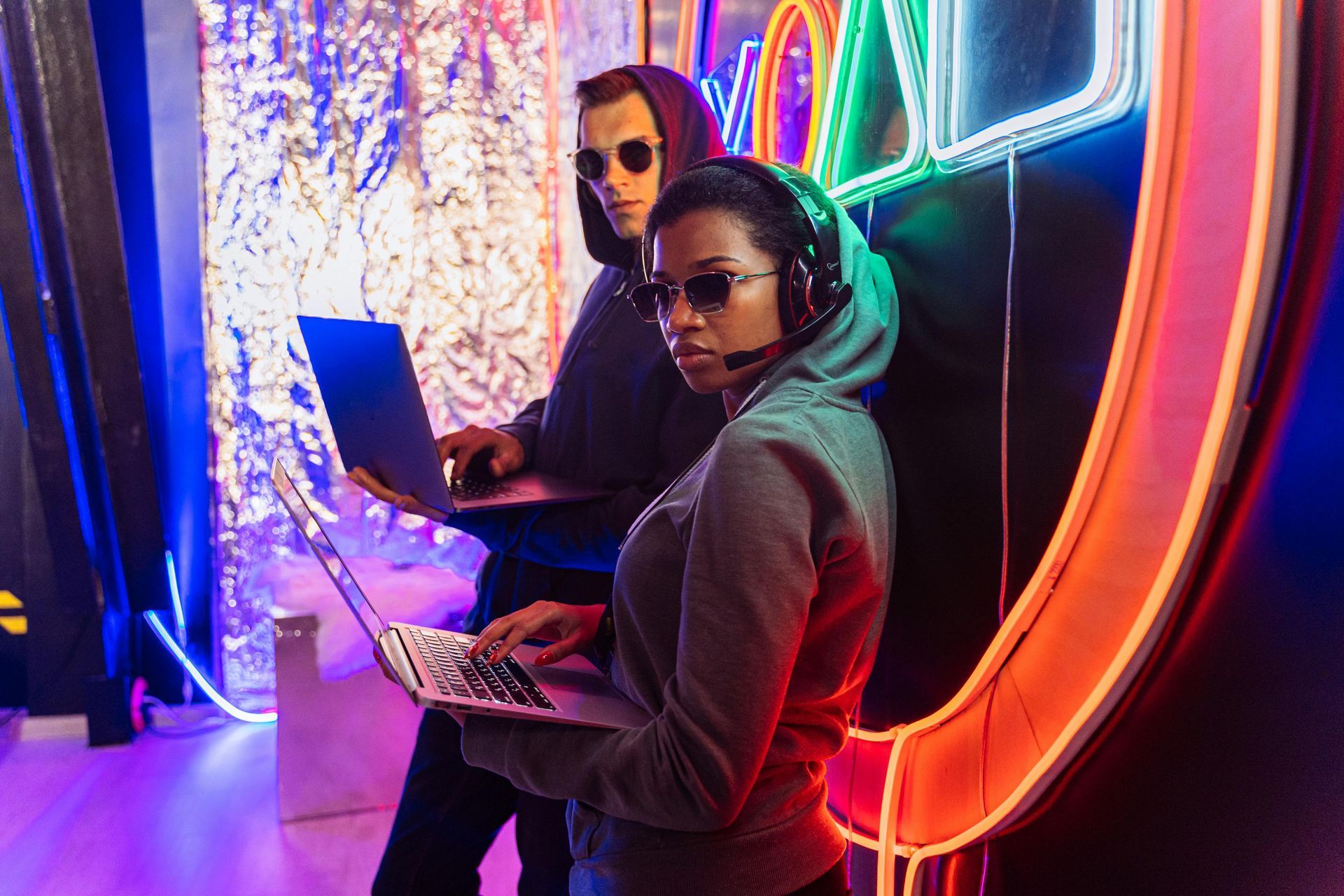 Two people in hoodies and sunglasses coding on laptops, neon lights in background.