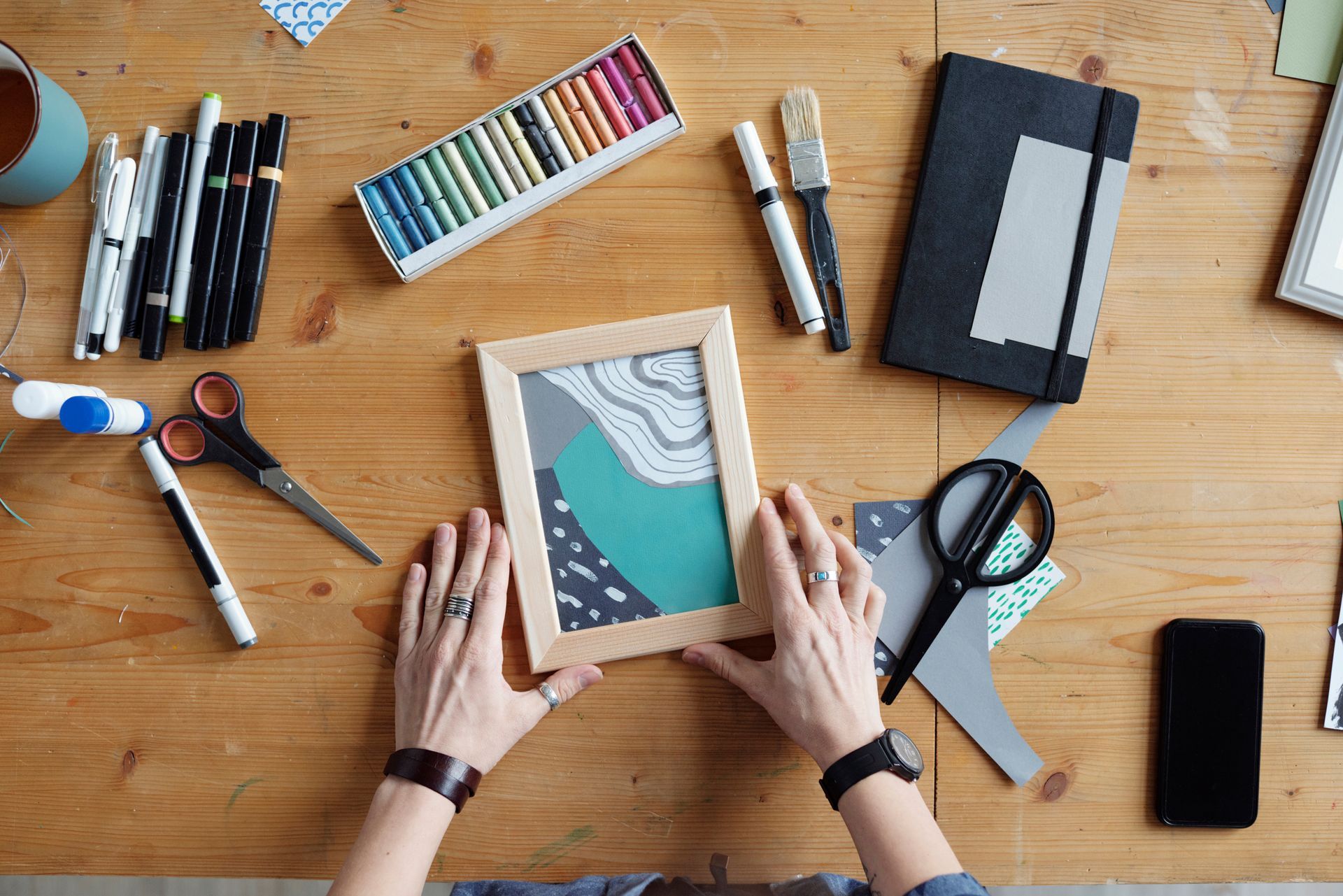 Hands arranging a framed art print on a desk with paints, scissors, notebook, and phone. Hands arranging a framed art print on a desk with paints, scissors, notebook, and phone.