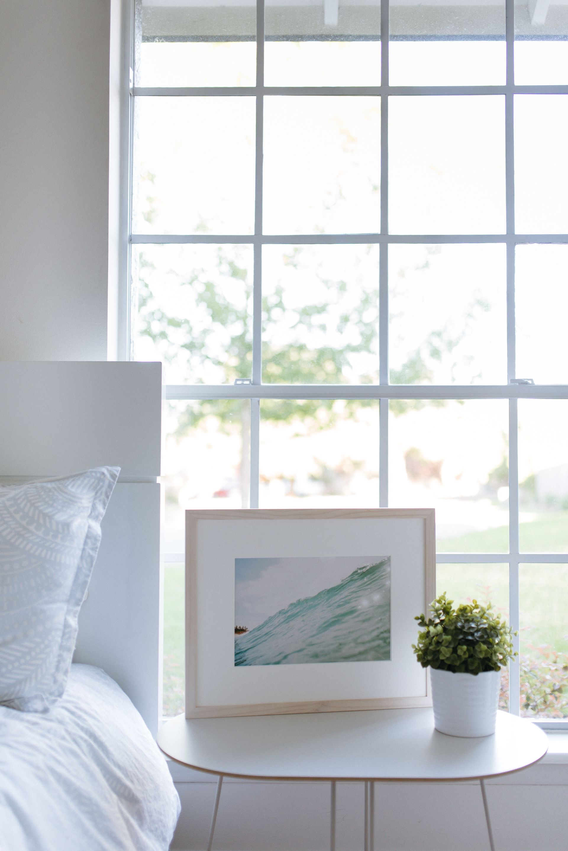 Cozy bedroom corner with a bed, round white table, framed art, and potted plant by a bright window Cozy bedroom corner with a bed, round white table, framed art, and potted plant by a bright window