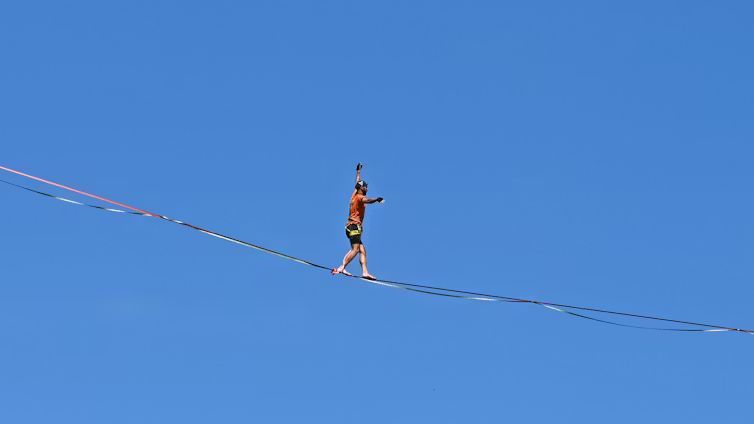 A man wearing an orange shirt balances carefully walks across a high slackline stretched against a solid, clear blue sky.