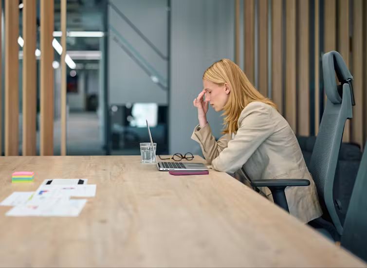 A blonde woman blazer sits at an office desk with a laptop, pinching the bridge of her nose with a stressed expression.