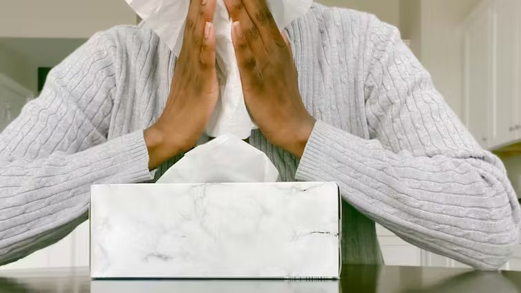 A person wearing a grey knit sweater holding a white tissue up to their face to blow their nose.