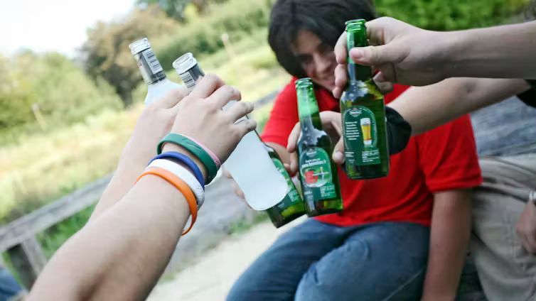 A close-up of several teenagers holding green glass beer bottles and a clear bottle together in a toast outdoors.