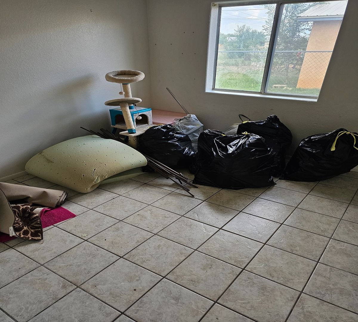 Spartan room with tiled floor, window, cat tree, green cushion, and several black trash bags along the wall