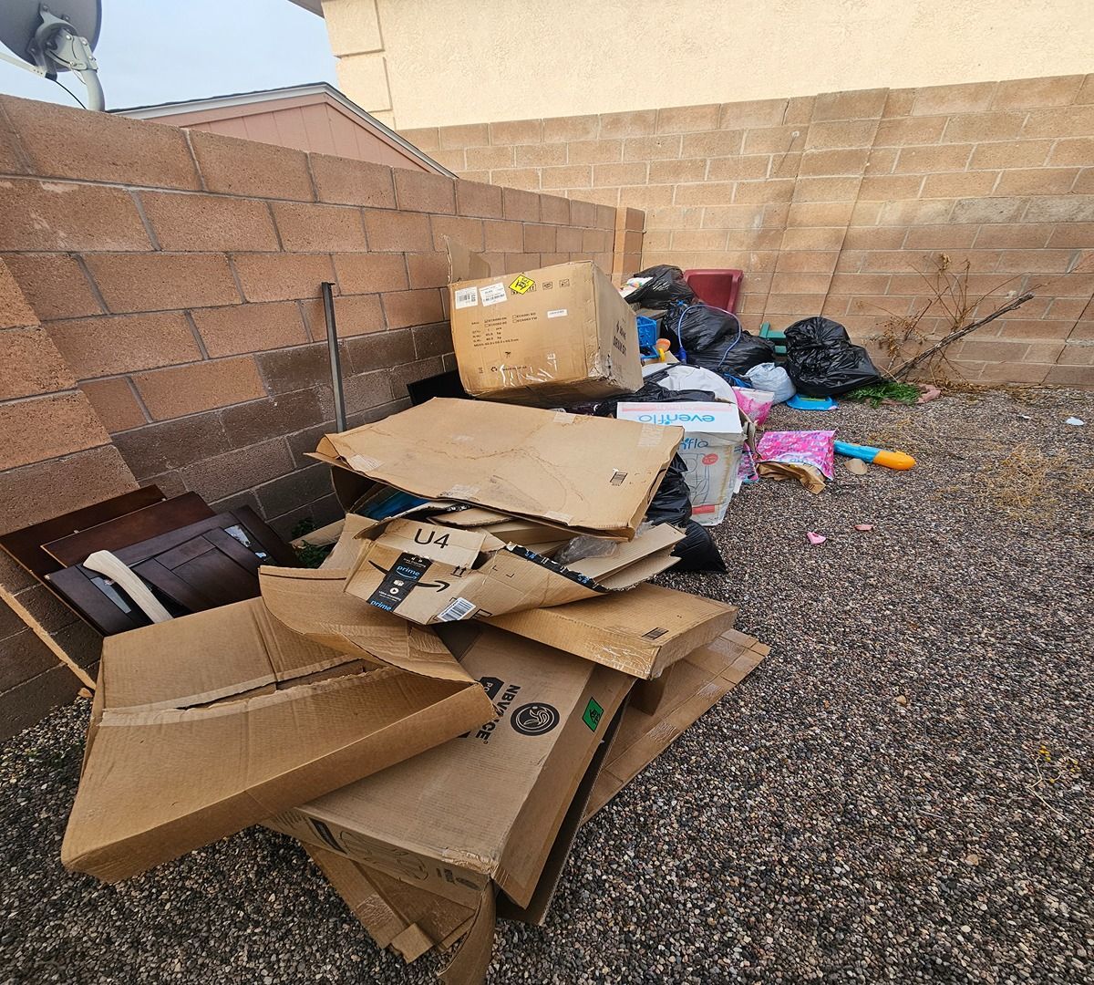 Cardboard and trash piled beside a tan block wall in a gravel yard.