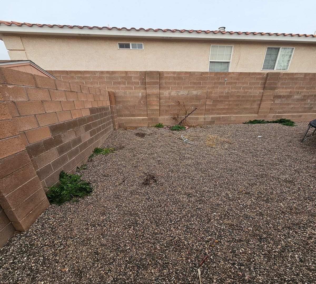 Empty gravel backyard with tan block walls and sparse weeds near a beige house.