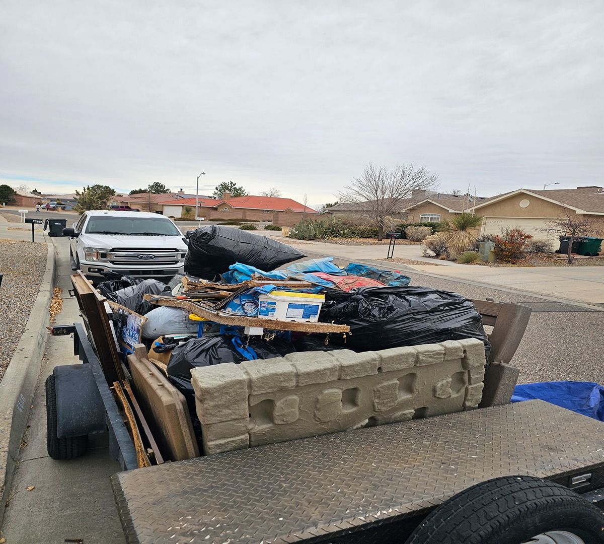 Trailer loaded with trash bags and debris on a suburban street behind a white pickup truck