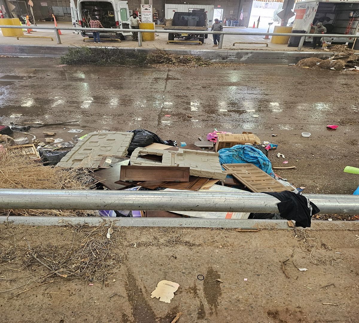 Trash-strewn muddy roadside with a metal barrier in the foreground and a wet street beyond.
