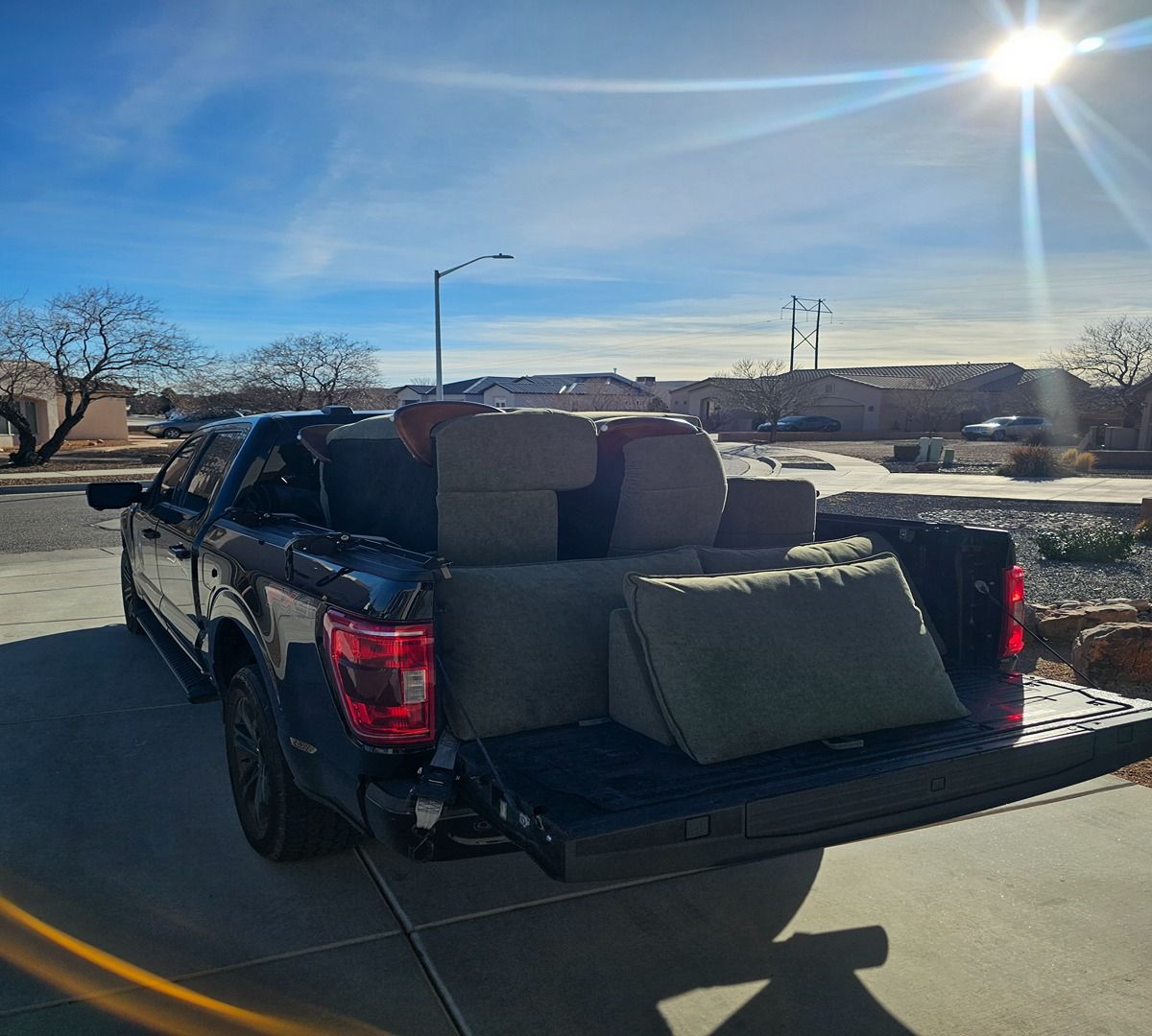 Black pickup truck with couch cushions and cardboard in the bed, parked in bright sunlight.