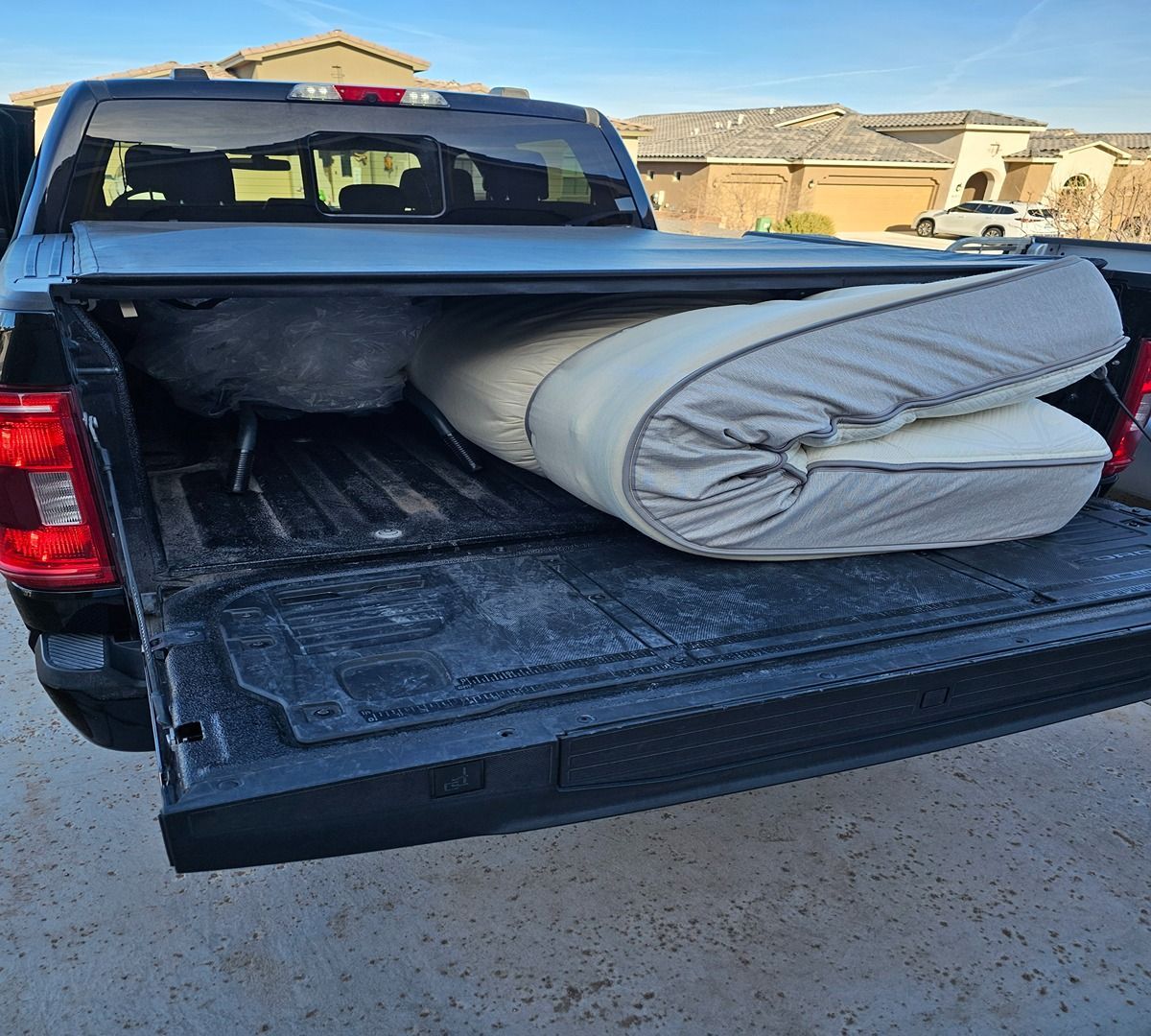 Pickup truck bed loaded with a rolled gray mattress on a driveway in a suburban neighborhood