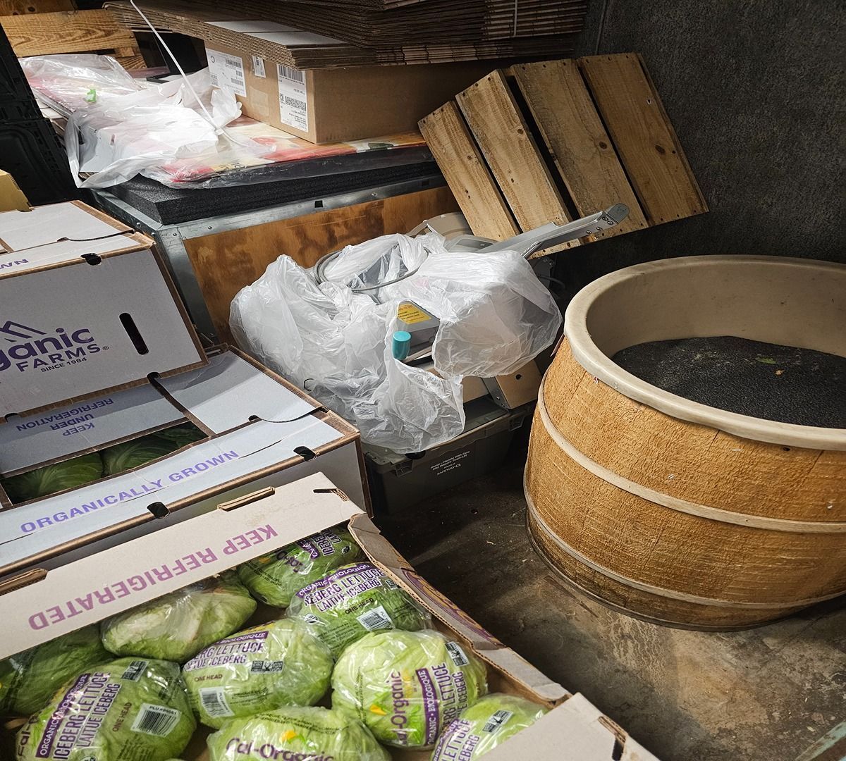 Grocer’s storage area with cardboard boxes, plastic bags, and a wooden bin of dark produce.