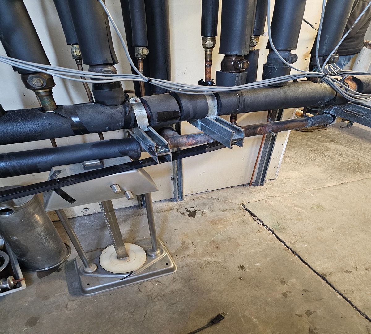Industrial pipes and valves along a wall above a concrete floor, with a support frame below