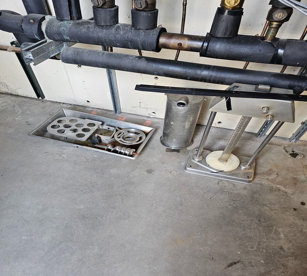 Industrial piping and floor-mounted valves above a concrete floor with drainage grates and a small metal bin