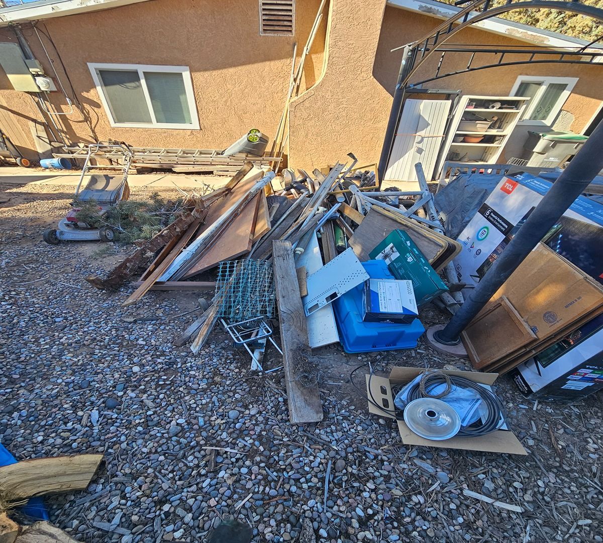 Debris and broken items piled in a gravel yard beside a brick house.