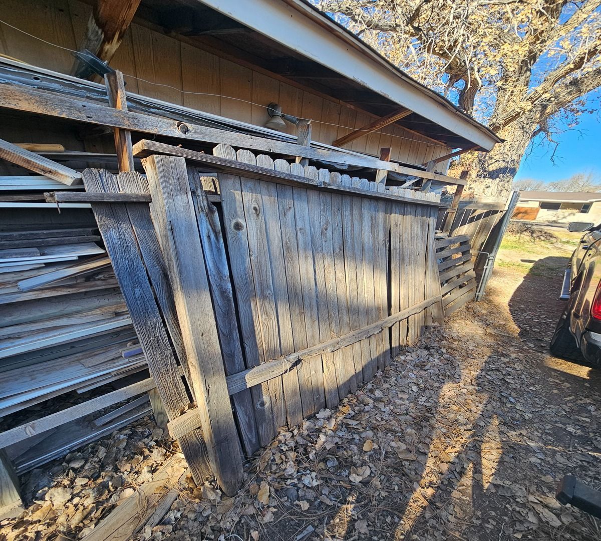 Weathered wooden gate leaning against a shed in a rustic yard with fallen leaves and long shadows