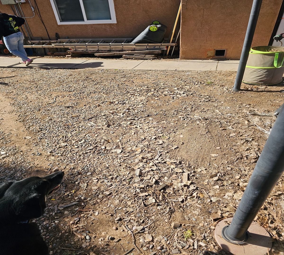 Rocky backyard work area with a brick house wall, window, pipe, and two workers near the edges.